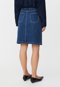 Jupe en jean bleu foncé, longueur au genou, avec poches arrière et ourlet surpiqué. Portée avec des chaussures noires sans lacets.