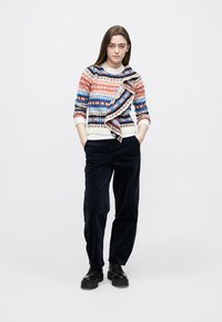 Camisola de malha colorida com padrões intrincados em laranja, azul e branco, combinada com calças de veludo cotelê pretas e botas pretas robustas.