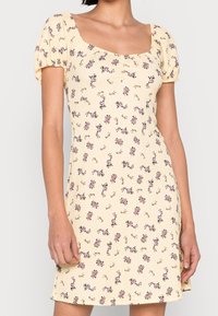 Robe florale jaune avec de courtes manches bouffantes, texture côtelée, décolleté carré et petits motifs de fleurs violettes partout.