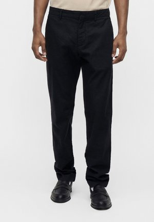 Homme portant un pantalon noir droit et des chaussures noires sans lacets sur un fond uni, montré de la taille aux pieds.