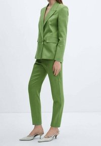 Blazer vert cintré et pantalon assorti en tissu texturé, avec des revers crantés et un bouton doré unique. Porté avec des escarpins slingback pointus.