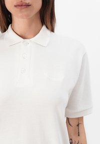 Valge polo särk, mis on valmistatud tekstureeritud kangast. Omab klassikalist kraed ja kolme nuppu. Vasakul rinnaosal on tagasihoidlik logo.