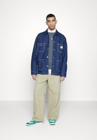Jeansjacka med fyra framfickor, blå färg, vit sömnad. Bärs över en grå tröja och krämfärgade byxor med gröna och blå sneakers.