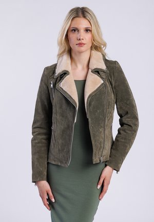 Žena oblečená v zelené semišové motocyklové bundě s krémovým límcem, sebevědomě stojící před jednobarevným pozadím.