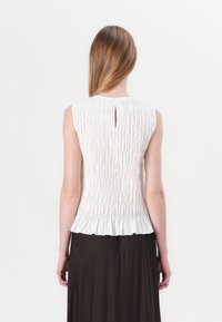 White sleeveless top with textured smocking, keyhole back detail, and a ruffled hem. Worn with a dark, flowing skirt.