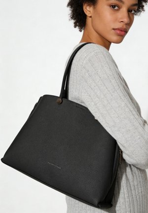 Femme portant un pull gris clair et un grand sac cabas en cuir noir sur l'épaule sur fond blanc.