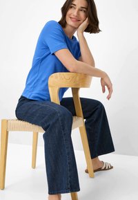 T-shirt di cotone blu con maniche corte abbinato a pantaloni larghi in denim scuro. Seduto su una sedia di legno con seduta intrecciata, indossa sandali bianchi.