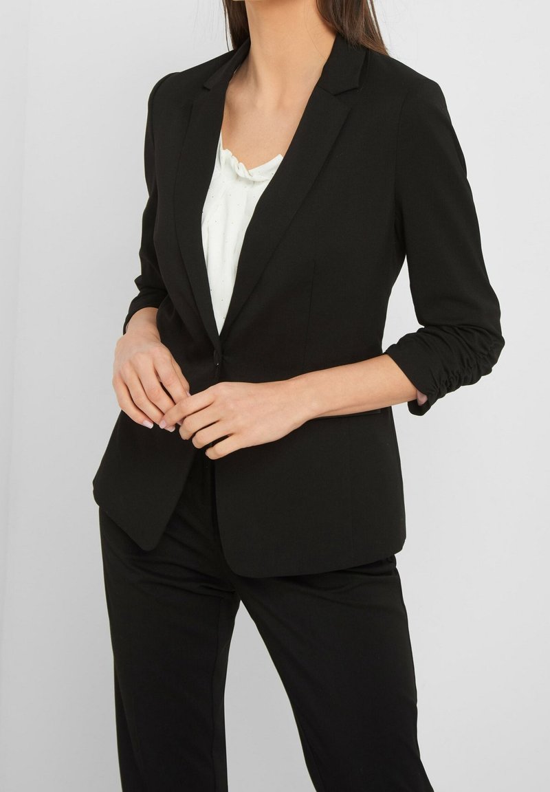 Femme portant un blazer noir cintré avec des manches froncées sur un haut blanc, associé à un pantalon noir, debout devant un fond uni.