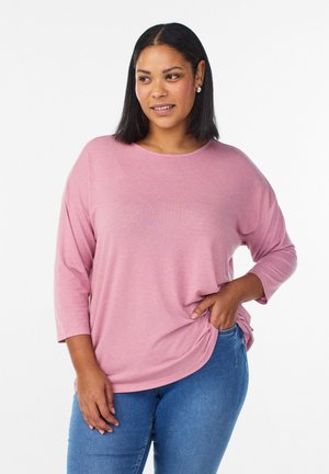 Žena v ružovej voľnej dlhorukávovej košeli a modrých džínsoch, stojaca s jednou rukou v vrecku na bielom pozadí.