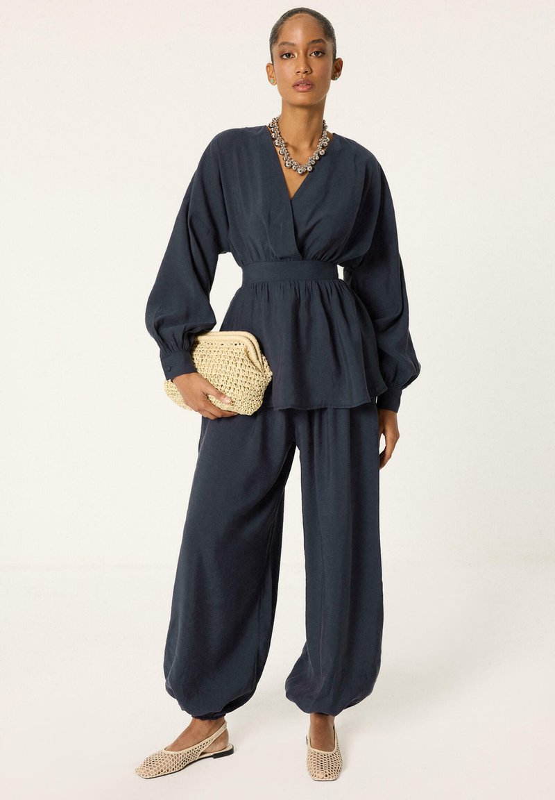 Femme portant un chemisier bleu marine à manches longues et un pantalon ample, tenant une pochette tissée beige, portant des ballerines en maille beige et un collier imposant.