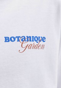 Άσπρο ύφασμα με το κείμενο «BOTanique» σε μπλε έντονα γράμματα και «Garden» σε καφέ γραφικό γράψιμο.