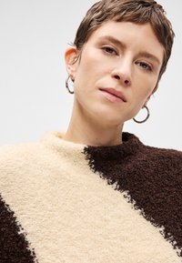 Πλεκτό πουλόβερ με διαγώνιο σχέδιο σε κρέμα και σκούρο καφέ χρώμα, με ψηλό λαιμό και μαλακό, χνουδωτό ύφασμα.