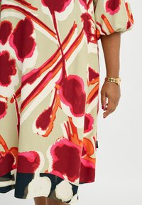 Kleid mit floralem Muster auf leicht beige Hintergrund. Enthält kräftige rote und pinke Blüten, orangefarbene Akzente und einen dunklen Saum. Kurze Puffärmel.