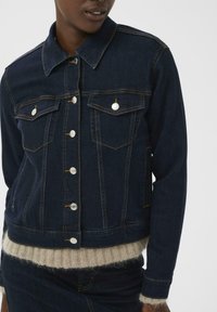 Dunkle Jeansjacke mit schmaler Passform, kontrastierenden gelben Nähten, Metallknöpfen und zwei Brusttaschen. Weicher Strickpullover ist am Saum sichtbar.