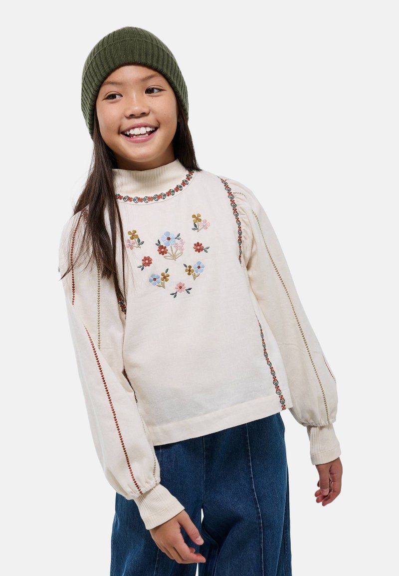 Pull crème avec broderie florale, col montant côtelé et manches bouffantes. Associé à un pantalon en jean bleu à jambes larges et un bonnet vert olive.