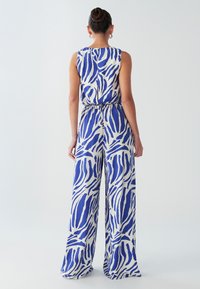 Plavi i bijeli kombinezon s prugama, širokih nogavica, opuštenog kroja i strukom prikupljenim u gumicu. Izrađen od lagane tkanine glatke teksture.