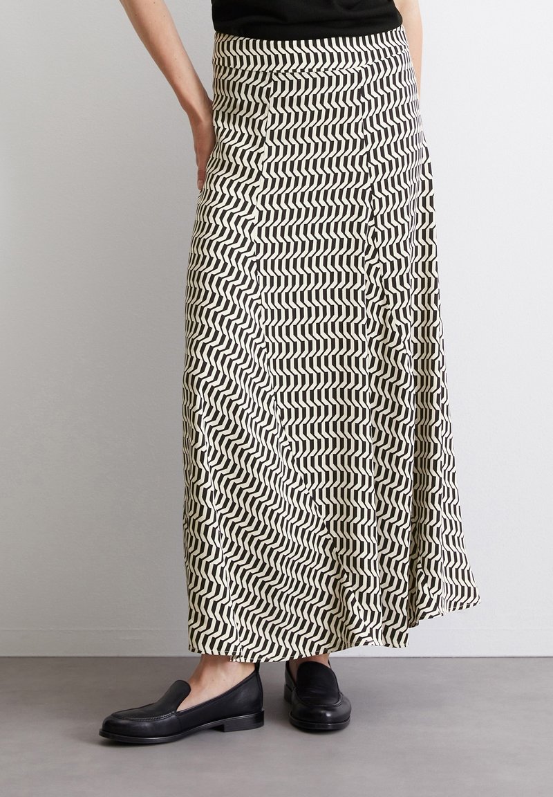 Personne portant une longue jupe noire et blanche à motif zigzag et des mocassins noirs, debout contre un mur blanc uni.