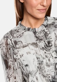 Durchsichtige, hellgraue Bluse mit einem Rüschenausschnitt. Verfügt über ein schwarzes Skizzenmotiv mit Gebäuden und Landschaften. Texturiert, fließender Stoff.