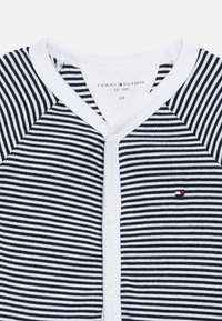 Λευκό και ναυτικό ριγέ βρεφικό φορμάκι με λευκά κουμπιά πάνας και μικρό κόκκινο, λευκό και ναυτικό λογότυπο Tommy Hilfiger στο στήθος.