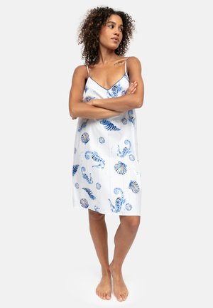 Femme aux cheveux bouclés portant une robe blanche sans manches avec des motifs de coquillages et d’hippocampes bleus et oranges, debout pieds nus, les bras croisés.