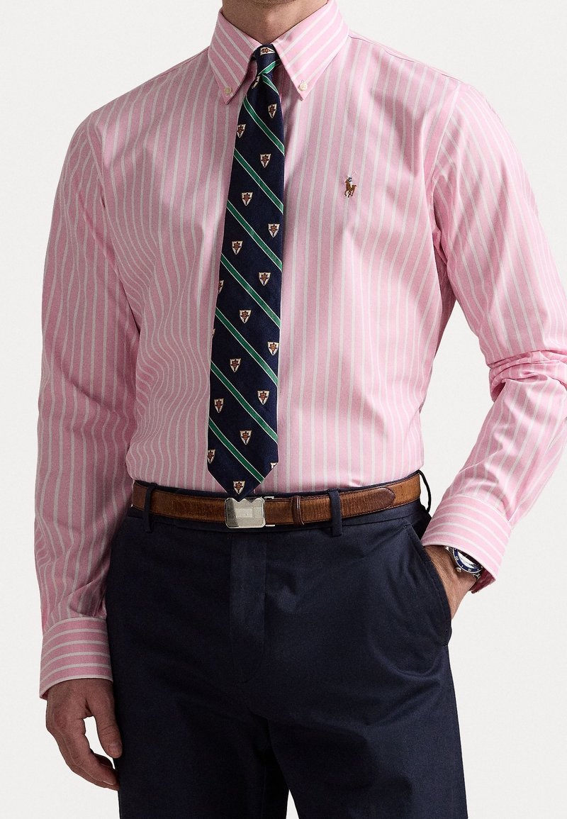 Pruhovaná růžová košile na knoflíky s námořnickou kravatou s vzorem emblémů a zelenými pruhy, doplněná tmavými kalhotami a hnědým páskem.