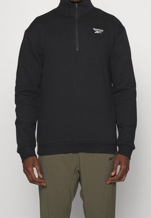 Czarna bluza Reebok z ćwiartkowym zamkiem, noszona przez osobę w oliwkowych spodniach Reebok, stojącą z rozluźnionymi rękami na jednolitym tle.