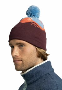 Gorro de punto en marrón con un pompón azul, con un diseño de montaña naranja y azul en la parte superior. Textura suave y ajuste ceñido.