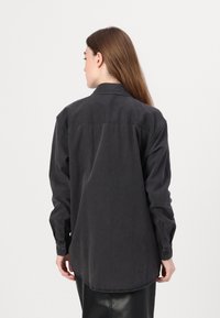 Melnas kokvilnas krekls ar brīvu piegriezumu, ar pogu aprocēm un muguras jostu dizainu. Gluda tekstūra un nedaudz izliekts apakšmalas dizains komfortam.