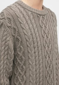 Nahaufnahme einer Person, die einen taupefarbenen Pullover mit Zopfmuster und ribbelndem Rundhalsausschnitt trägt.