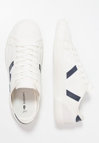 Valkoiset kangaskengät tummansinisin yksityiskohdin, joissa on nauhakiristyksellinen muotoilu, kumipohja ja pieni logo sivussa.