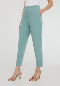 Pantaloni tapered verde menta con una texture liscia, chiusura frontale con bottone e tasche laterali, abbinati a sandali con tacco beige.