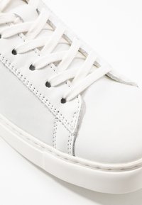 Sneaker in pelle bianca con stringhe piatte bianche, dettagli di cucitura sottili, texture liscia e suola in gomma dal design pulito.