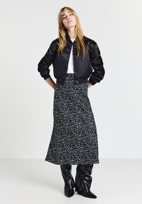 Jeune femme portant une veste bomber en satin noir, un haut blanc, une jupe midi à motifs noir et blanc, et des bottes à talons noires lâches, se tenant les mains sur les hanches.