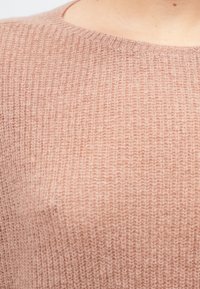 Svetle růžový pletený svetr s žebrovanou texturou a kulatým výstřihem, představující jemnou, načechranou tkaninu a jemnou barevnou variaci.