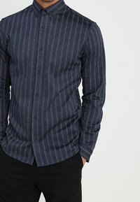 Man som bär en marinblå skjorta med knappar och vertikala röda och vita ränder, med händerna i fickorna på svarta byxor, mot en enfärgad ljus bakgrund.