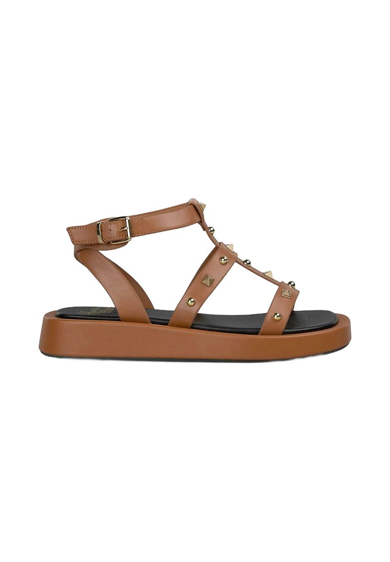 Sandal in sintetico marrone con cinturini e suola piatta nera, caratterizzata da borchie dorate e una chiusura con fibbia per la regolazione.