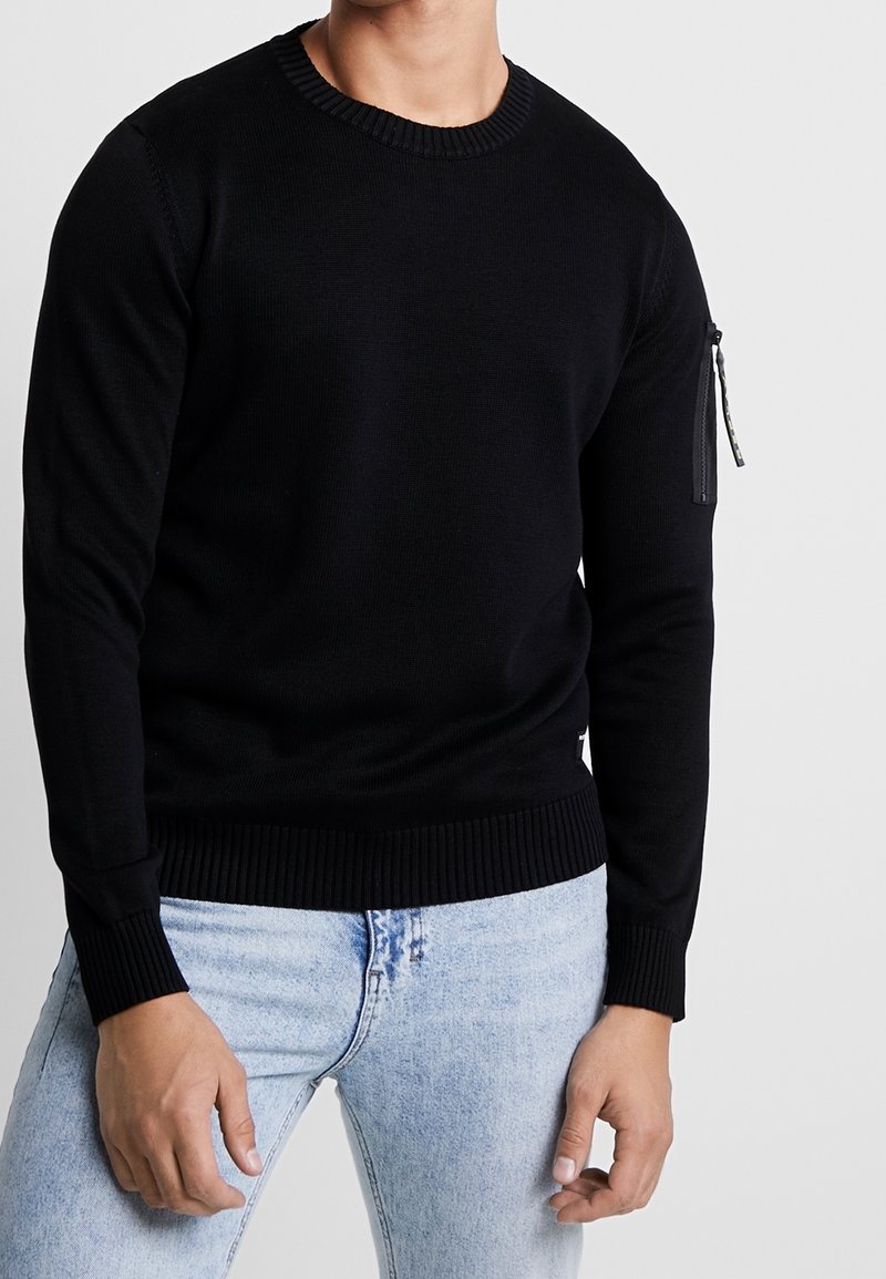 Homme portant un pull noir à manches longues avec une poche zippée sur la partie supérieure de la manche gauche et un jean bleu clair sur fond gris.