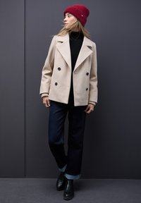 Abrigo de lana beige de doble botonadura, cuello alto negro, vaqueros anchos de mezclilla oscura y botines negros, complementados con un gorro de punto rojo.