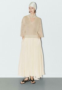 Vrouw in een beige gebreide trui met uitsnijding bij de kraag, lange crèmekleurige rok, zwarte sandalen en een witte hoofddoek, staande tegen een vlakke muur.