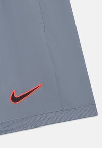 Shorts de sport gris avec un ourlet texturé. Présente un logo Nike noir entouré d'orange vif sur le côté gauche. Texture de tissu lisse.