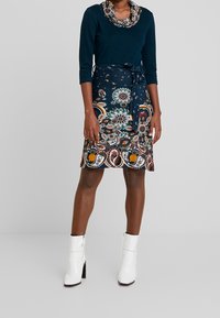 Långärmad grönblå topp med krage, i kombination med en mönstrad kjol som har blommönster i marinblått, orange och vitt. Vita ankelskor.