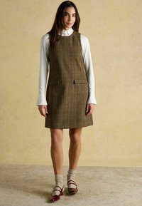 Braun-kariertes ärmelloses Kleid mit geradem Schnitt, ausgestattet mit zwei vorderen Taschen. Trägt man über einem langärmligen weißen Hemd und kombiniert mit weinroten Schuhen.