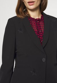 Černý blazer se sekvencovými hvězdnými akcenty, s pohodlným střihem, klopami a jednobarevným zapínáním na knoflík, přes červenou vzorovanou blůzu.