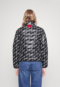 Sort pufferjakke med hvid tekstmønster, der prydes med "HUGO." Afsluttet med et rødt logo-tag på ryggen og et højt kravedesign.