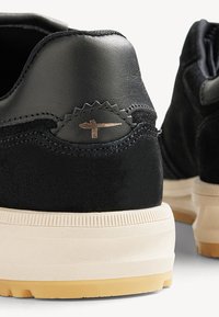 Black sneaker made of leather and suede, featuring a textured heel, light-colored sole, and a small metallic logo accent.