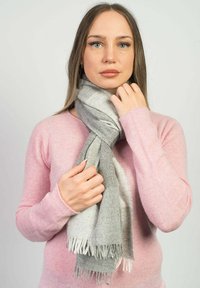 Sciarpa a scacchi grigia e bianca realizzata in materiale morbido con bordi frangiati, indossata sopra un maglione rosa chiaro da una persona che la tiene vicino al collo.