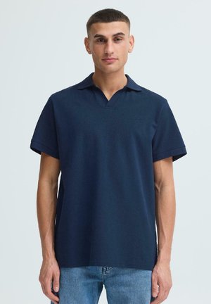 Jeune homme debout portant un polo bleu marine à manches courtes et un jean bleu clair, devant un fond uni clair.