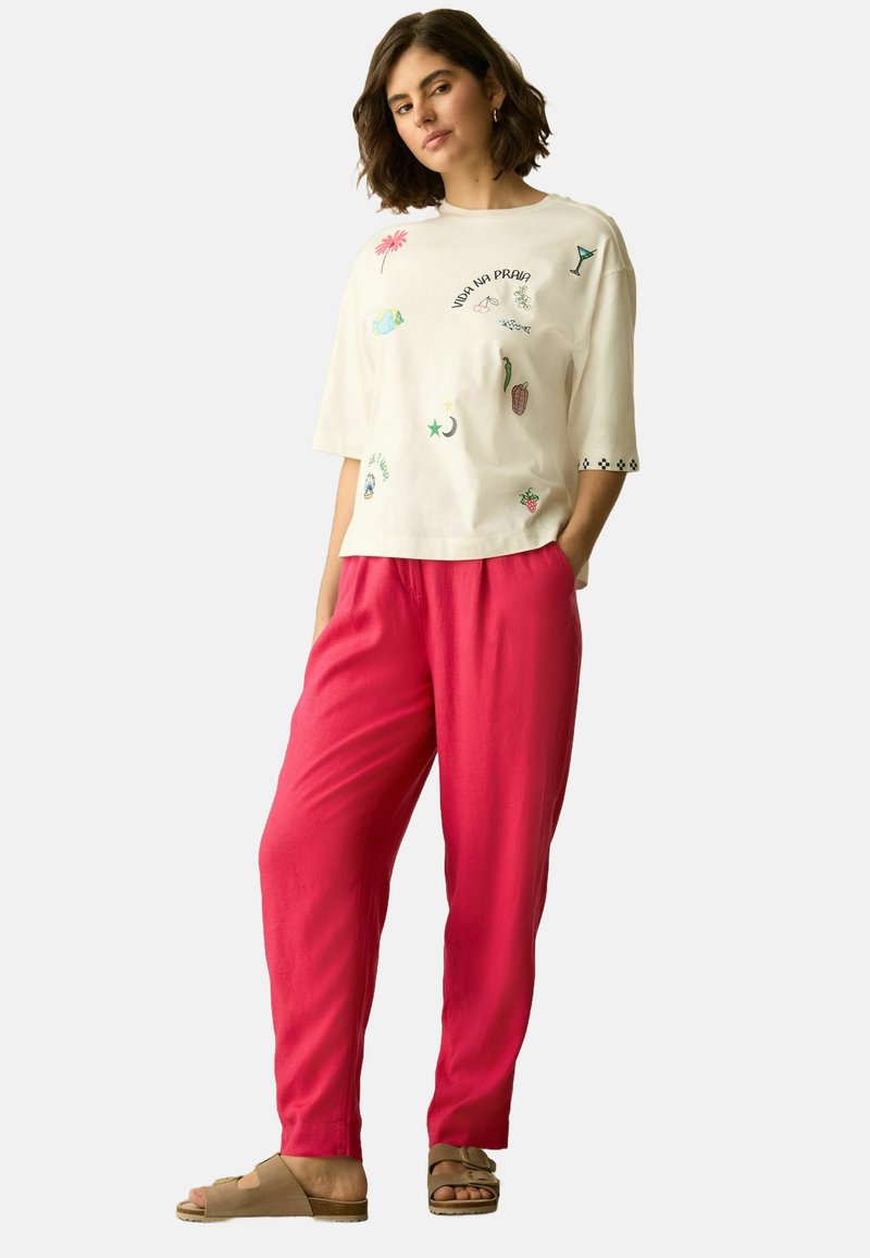 T-shirt en coton blanc avec des motifs colorés brodés, associé à un pantalon ample rose vif et des sandales marron clair.