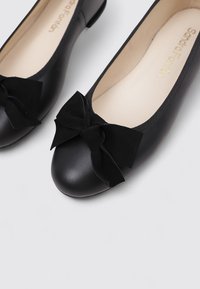 Bailarinas de cuero negro con punta redonda, que presentan un lazo de tela negra texturizada en la parte delantera y un interior suave en beige.