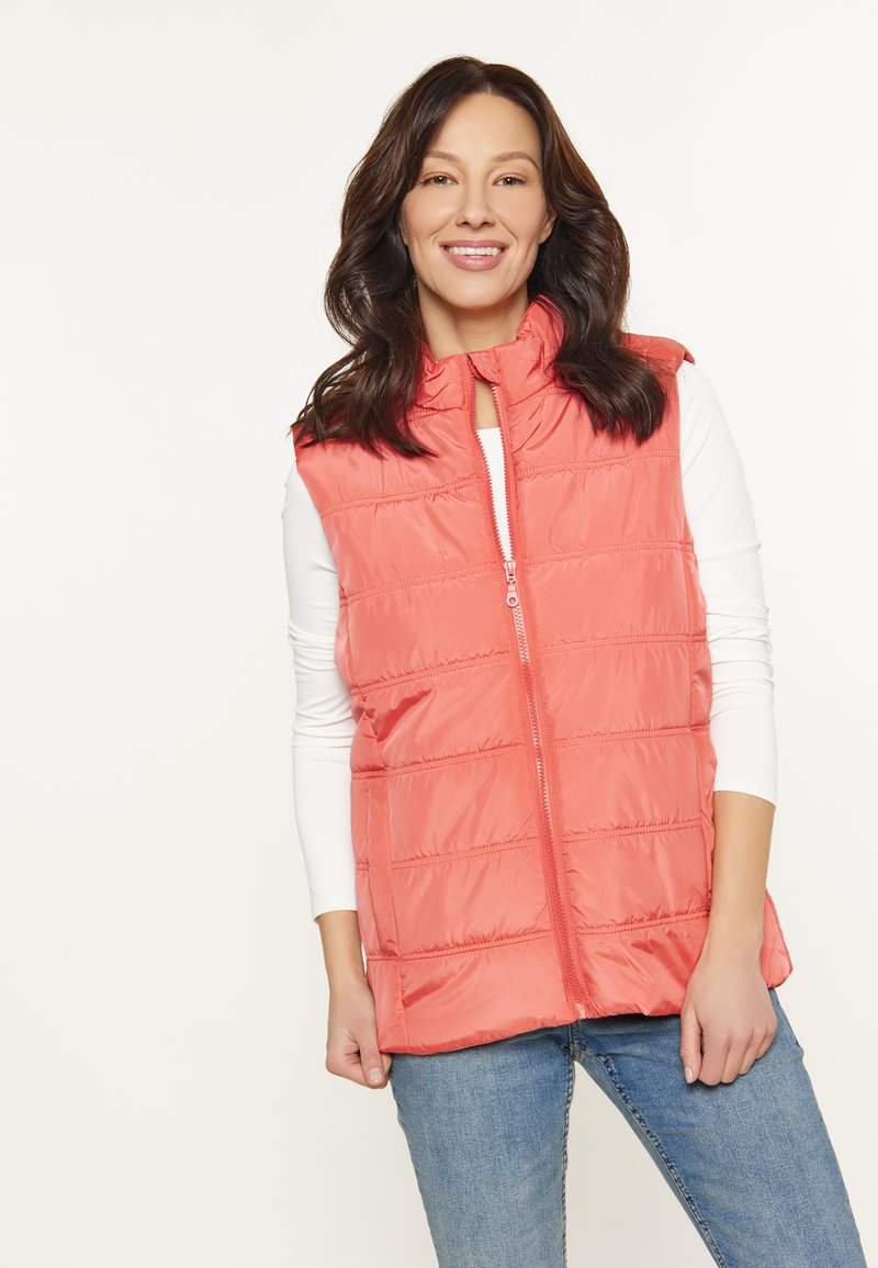 Koraal kleurige puffer vest met een hoge kraag, verticale naden en een rits aan de voorkant. Draag het over een wit longsleeve shirt, gecombineerd met blauwe jeans.